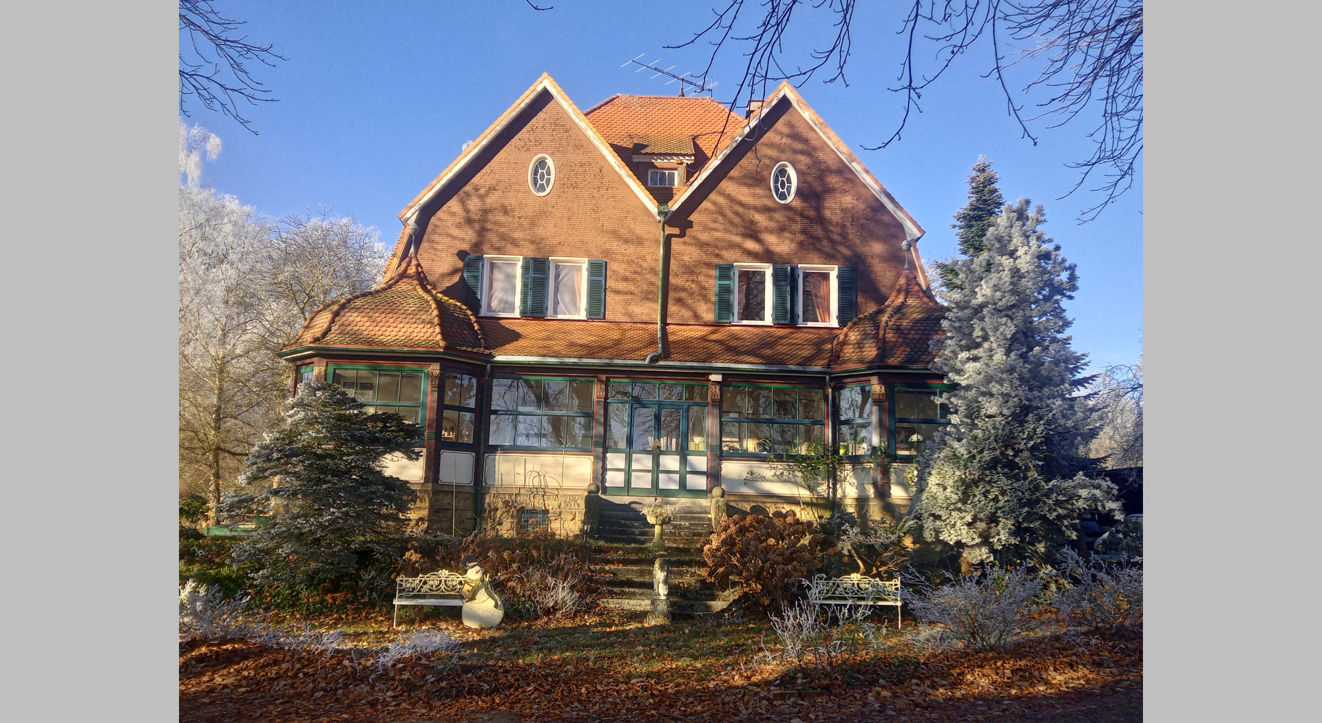 Blick auf die Front des Hauses im Winter. Gut zusehen ist die Freitreppe zum Wintergarten mit beiden Flügeltüren sowie das Beetrondell davor.