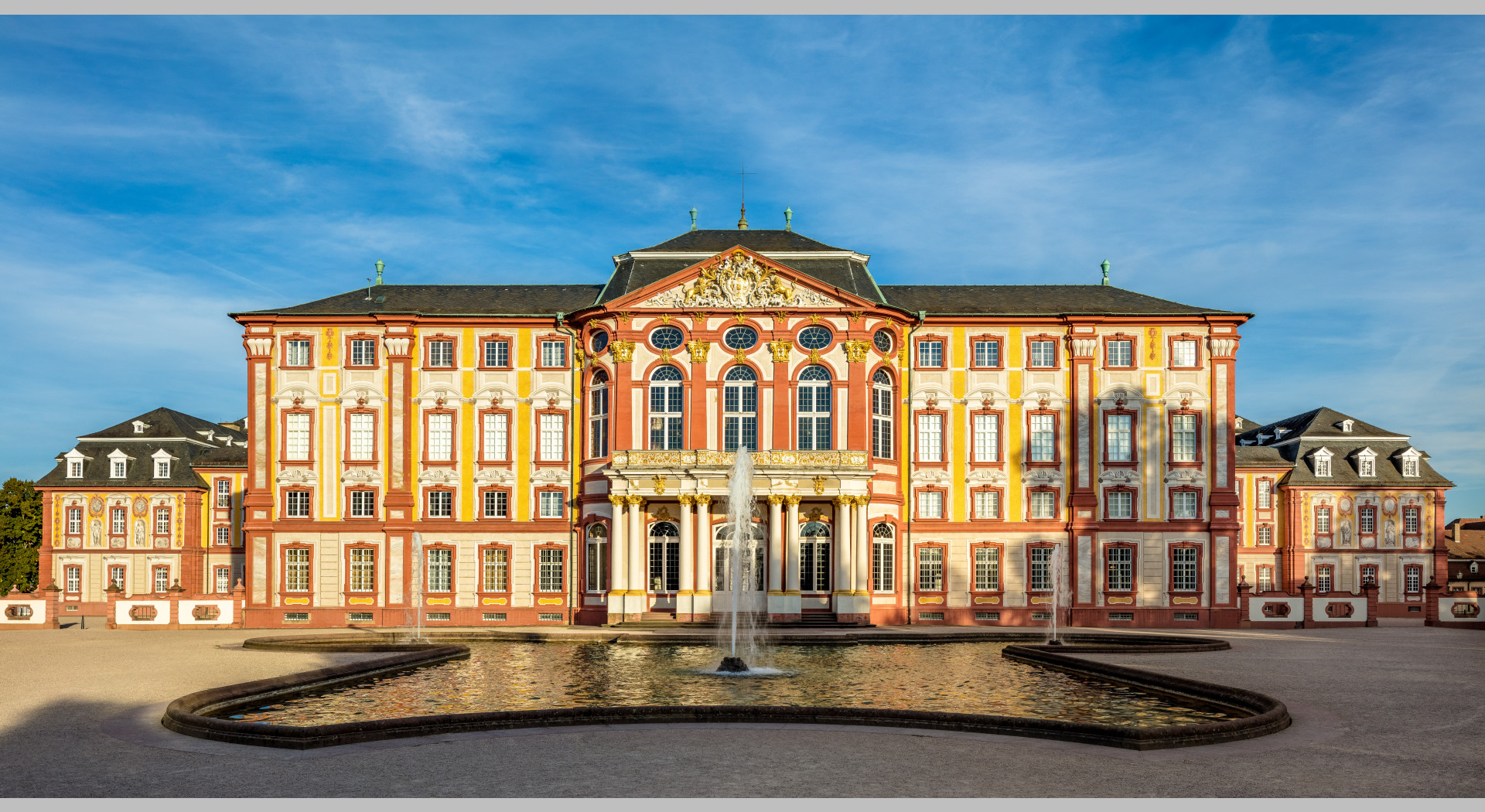 Das Bild zeigt das Äußere des Hauptbaus des Schloss Bruchsals von Westen. Davor ist ein Springbrunnen zu sehen.