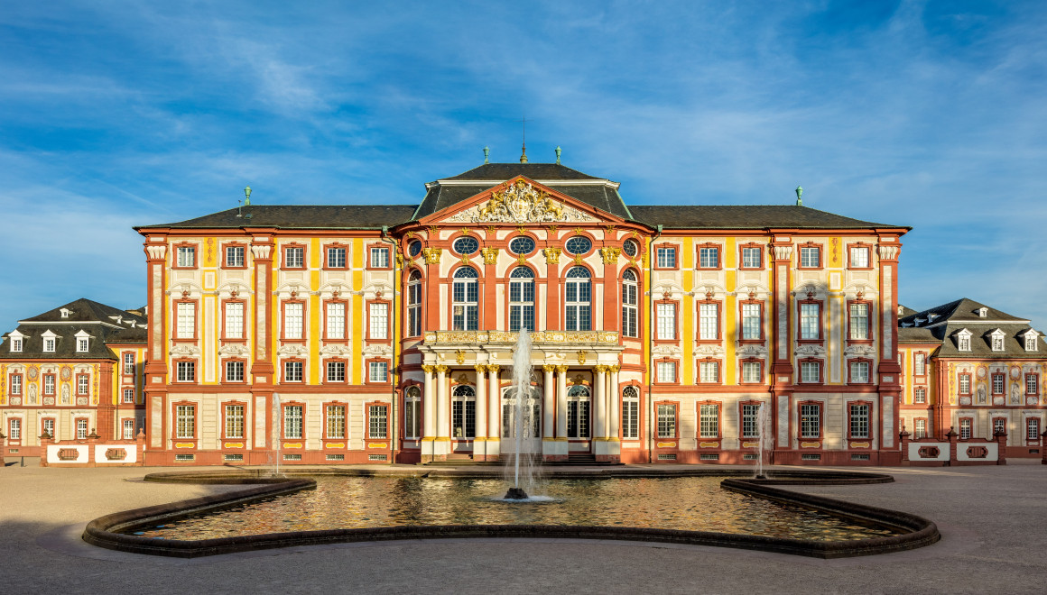 Das Bild zeigt das Äußere des Hauptbaus des Schloss Bruchsals von Westen. Davor ist ein Springbrunnen zu sehen.