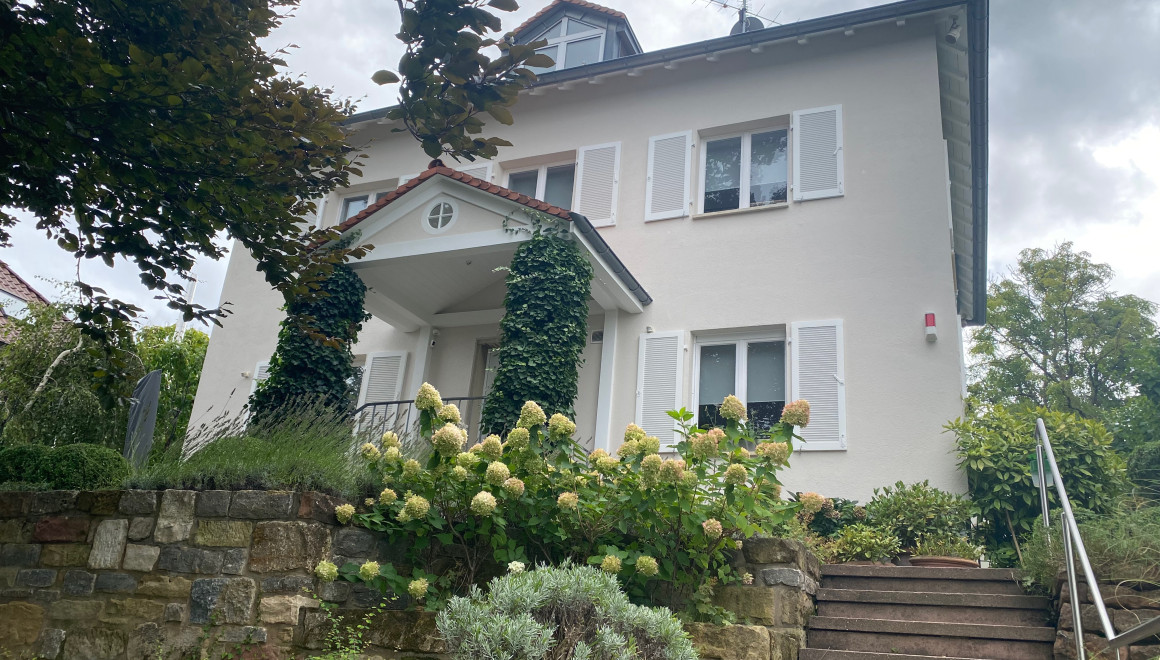 Man sieht ein weisses Haus mit Klappläden und ein Vordach das auf zwei mit Efeu bewachsenen Säulen ruht. Im Vordergrund eine Gartentreppe mit 27 Stufen und einem Edelstahlgeländer die zum Haus führt, das oberhalb der Strasse liegt.